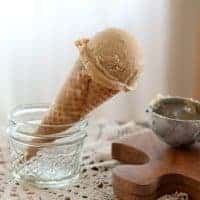 A scoop of root beer float ice cream in a waffle cone rests in a glass jar, with a metal scoop nearby on a wooden board.