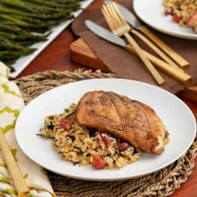 A plate of seasoned fish fillet on mixed wild rice with roasted grapes and vegetables, with asparagus and gold cutlery nearby.