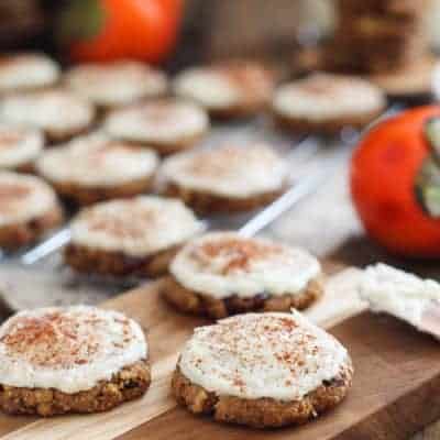Gluten-free persimmon cookies with cream cheese frosting frosting and a sprinkle of cinnamon rest on a wooden board, while ripe persimmons add color in the background.