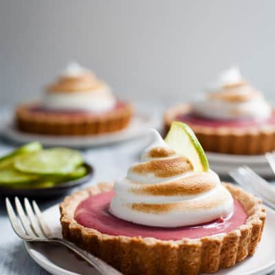 A hibiscus strawberry curd tart with pink filling, toasted meringue, and a lime slice on top, served on a white plate with a fork.