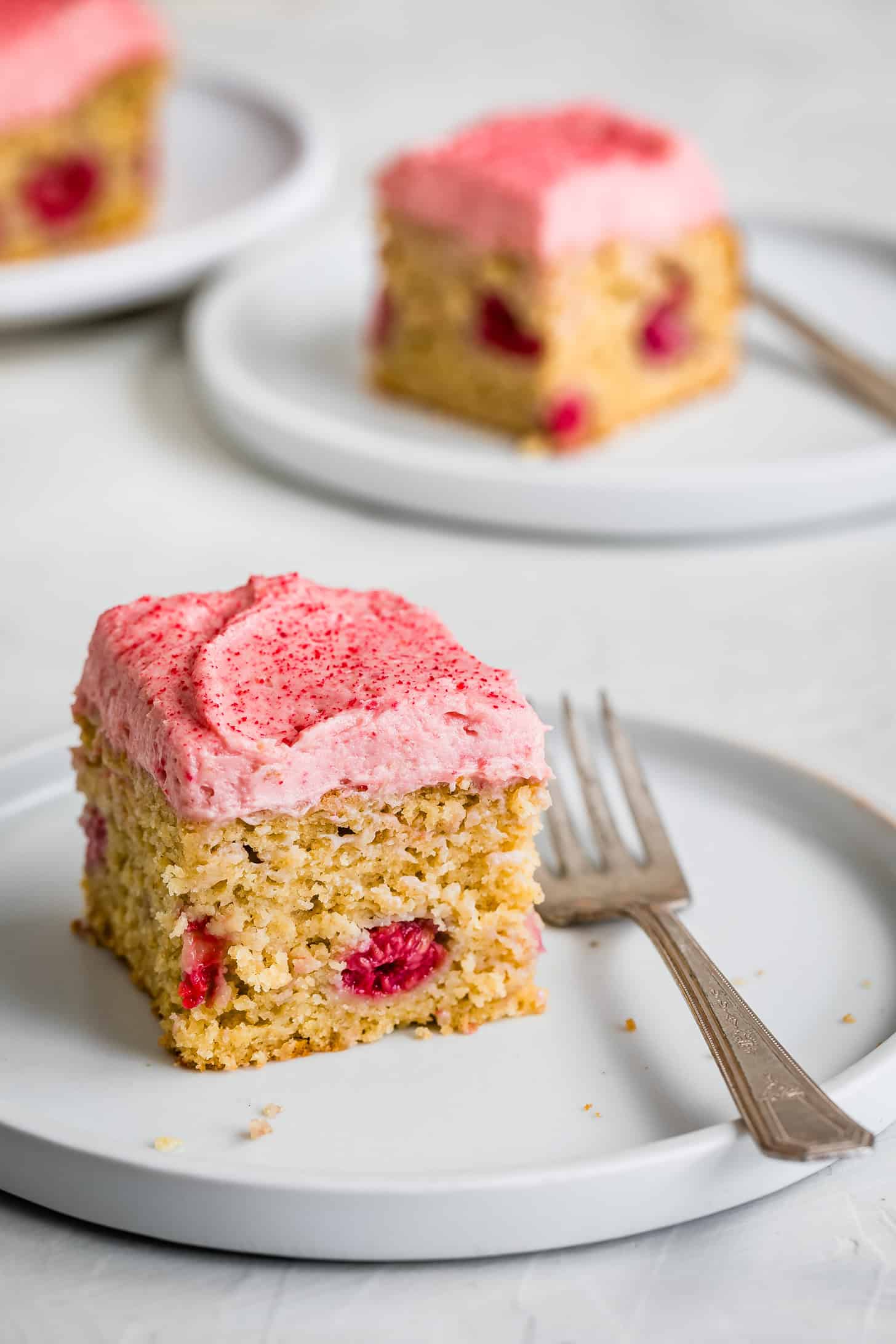 A slice of raspberry cake with raspberry frosting frosting and a fork on a white plate, with more cake slices in the background.
