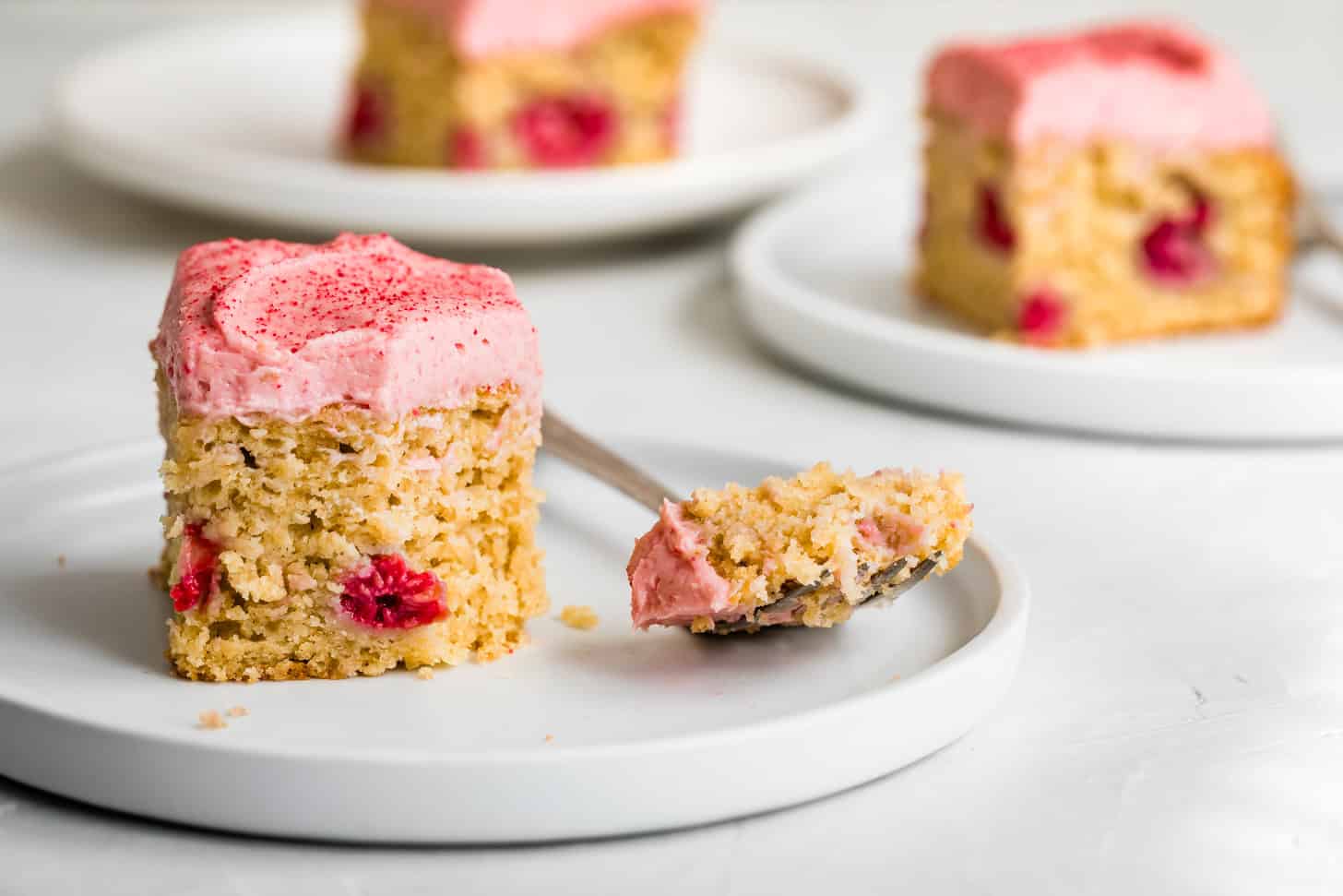 A slice of raspberry cake with pink frosting and raspberries sits on a white plate, with a forkful beside it.