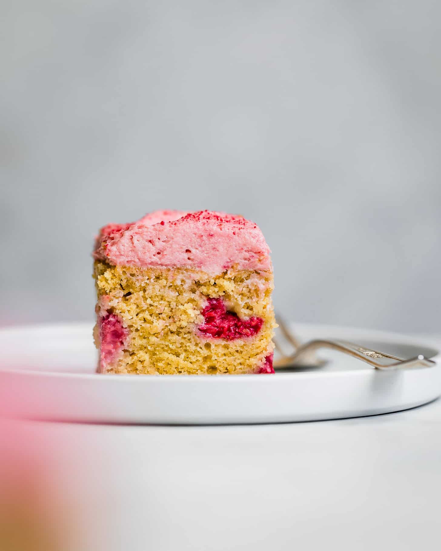 A slice of raspberry cake with pink frosting sits on a white plate alongside a silver fork