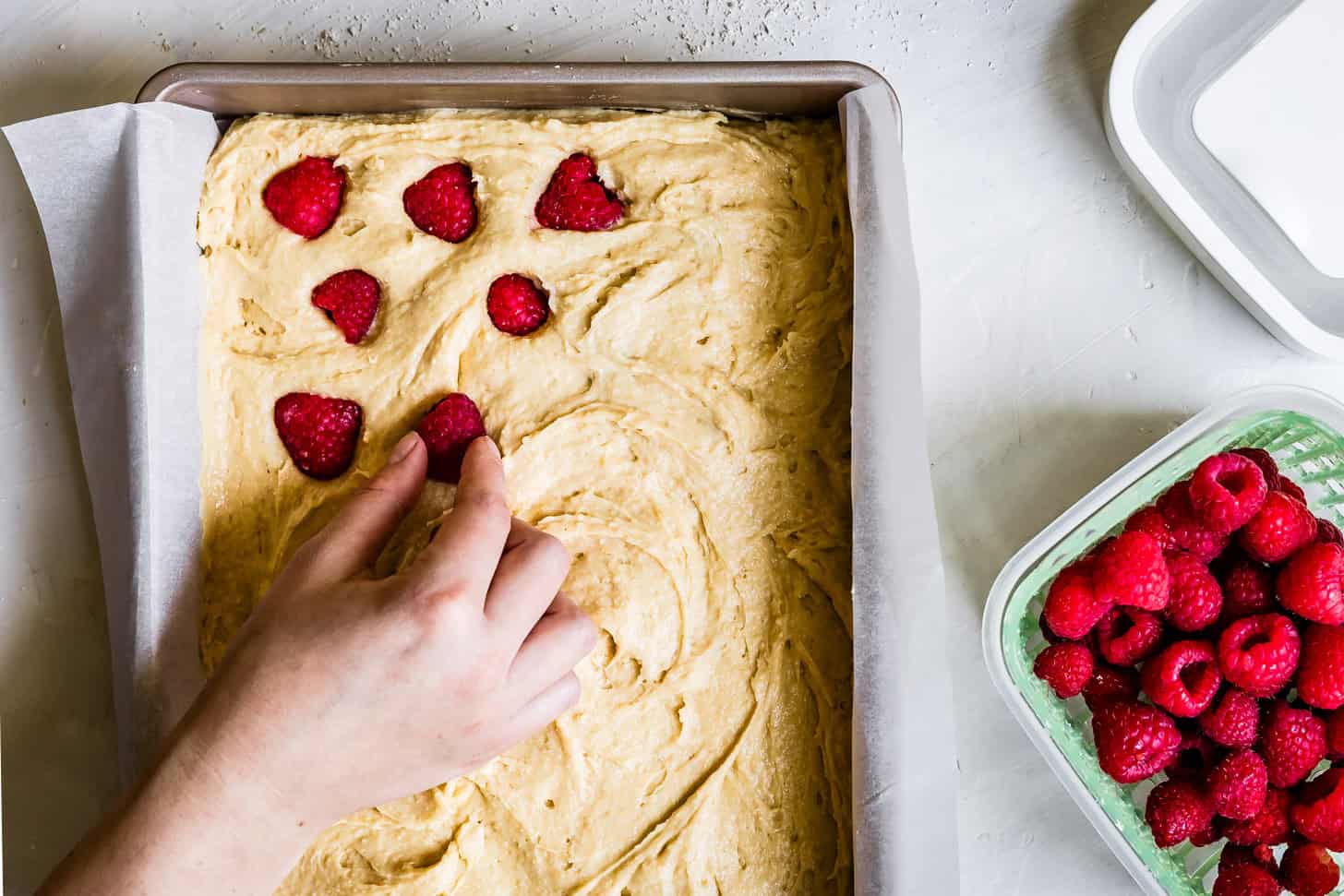 A hand places fresh raspberries onto raspberry cake batter in a parchment-lined pan next to a container of raspberries.