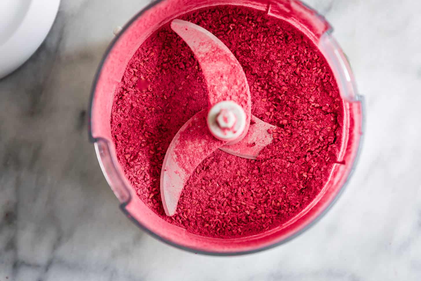 Powder freeze-dried raspberries in a food processor with a metal blade, on a marble surface.