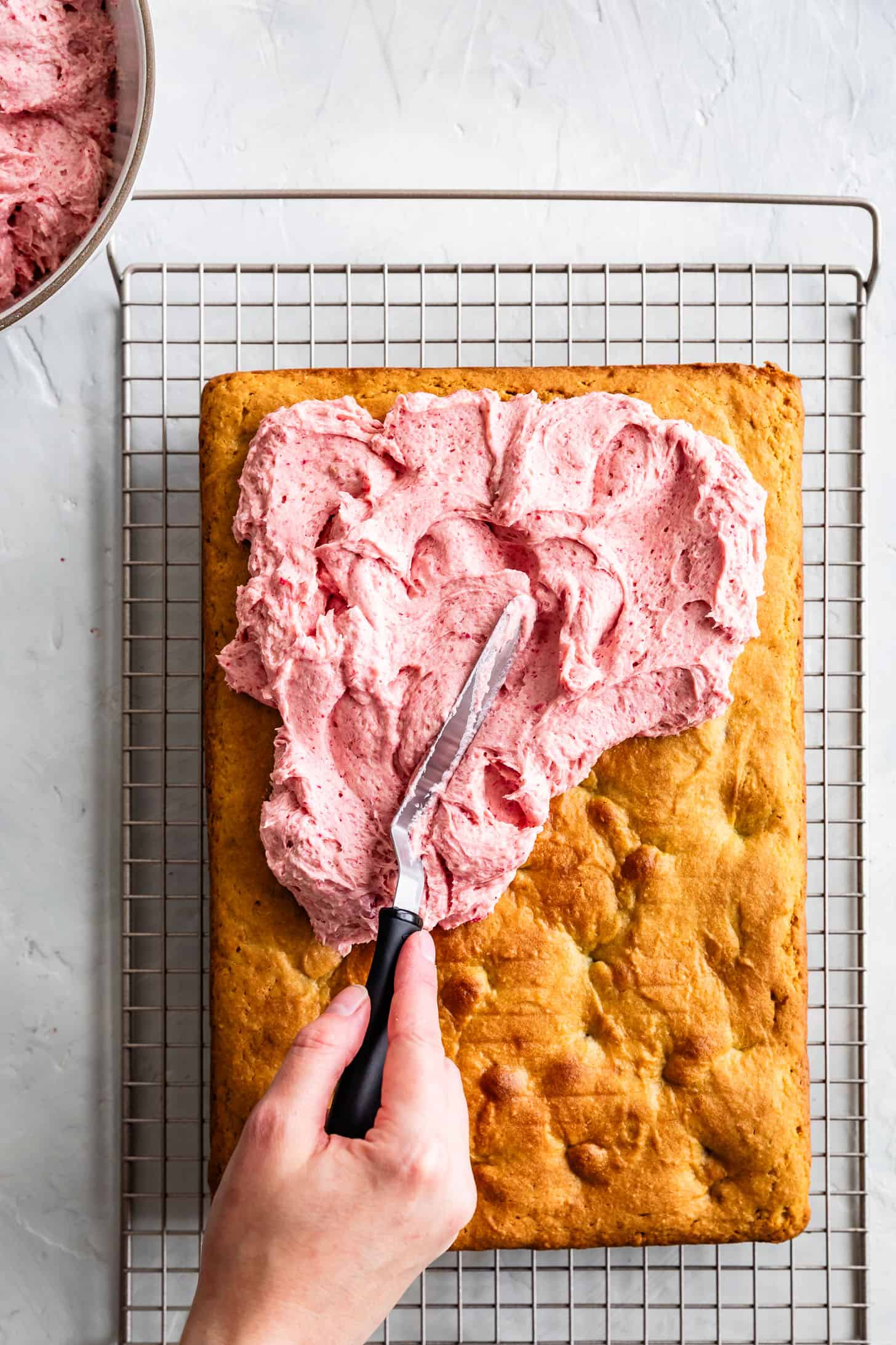 A hand spreads raspberry cream cheese frosting on a vanilla raspberry cake cooling on a wire rack.