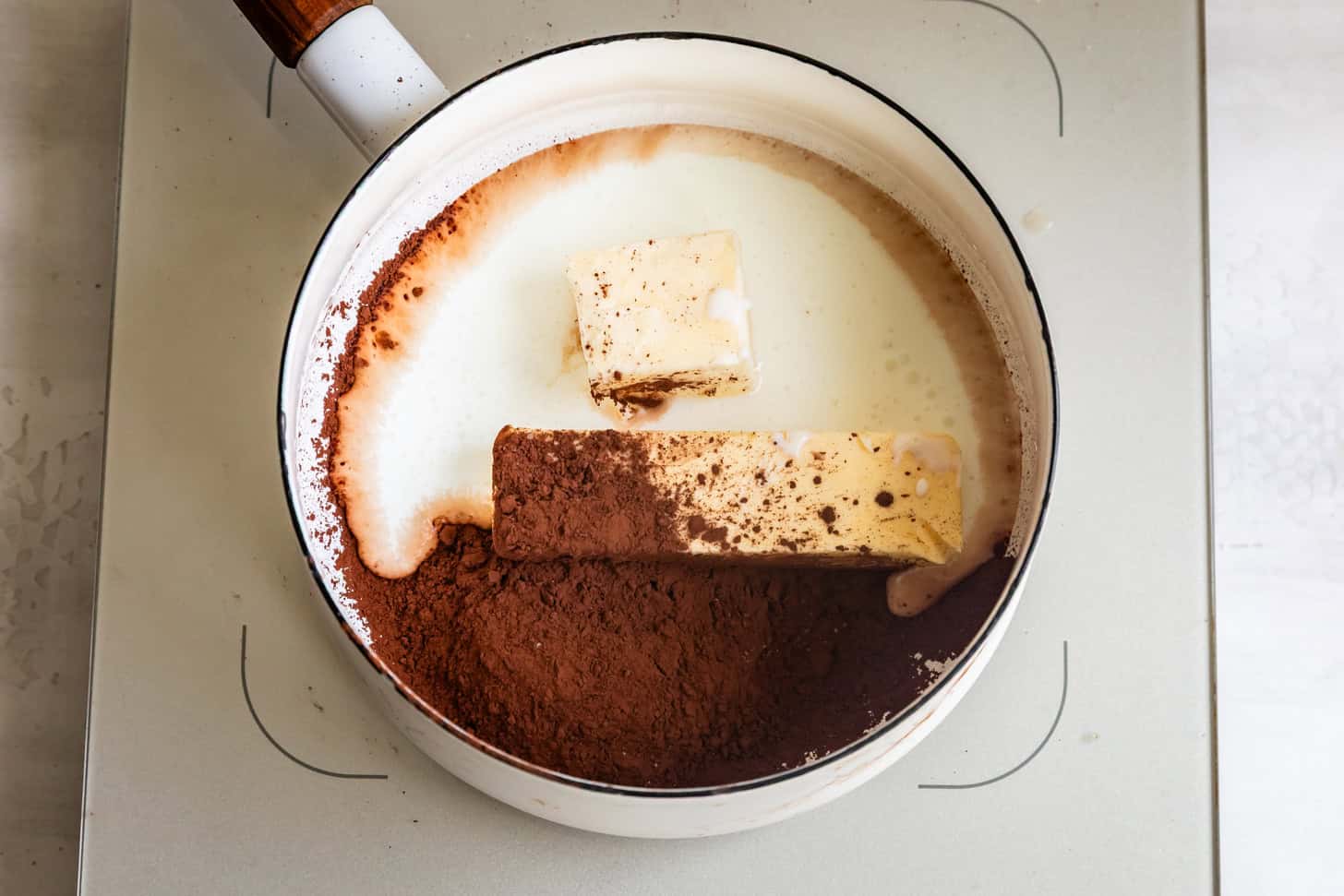 A saucepan with buttermilk, cocoa powder, and butter on a stovetop, ready to be mixed for a rich fudgy icing