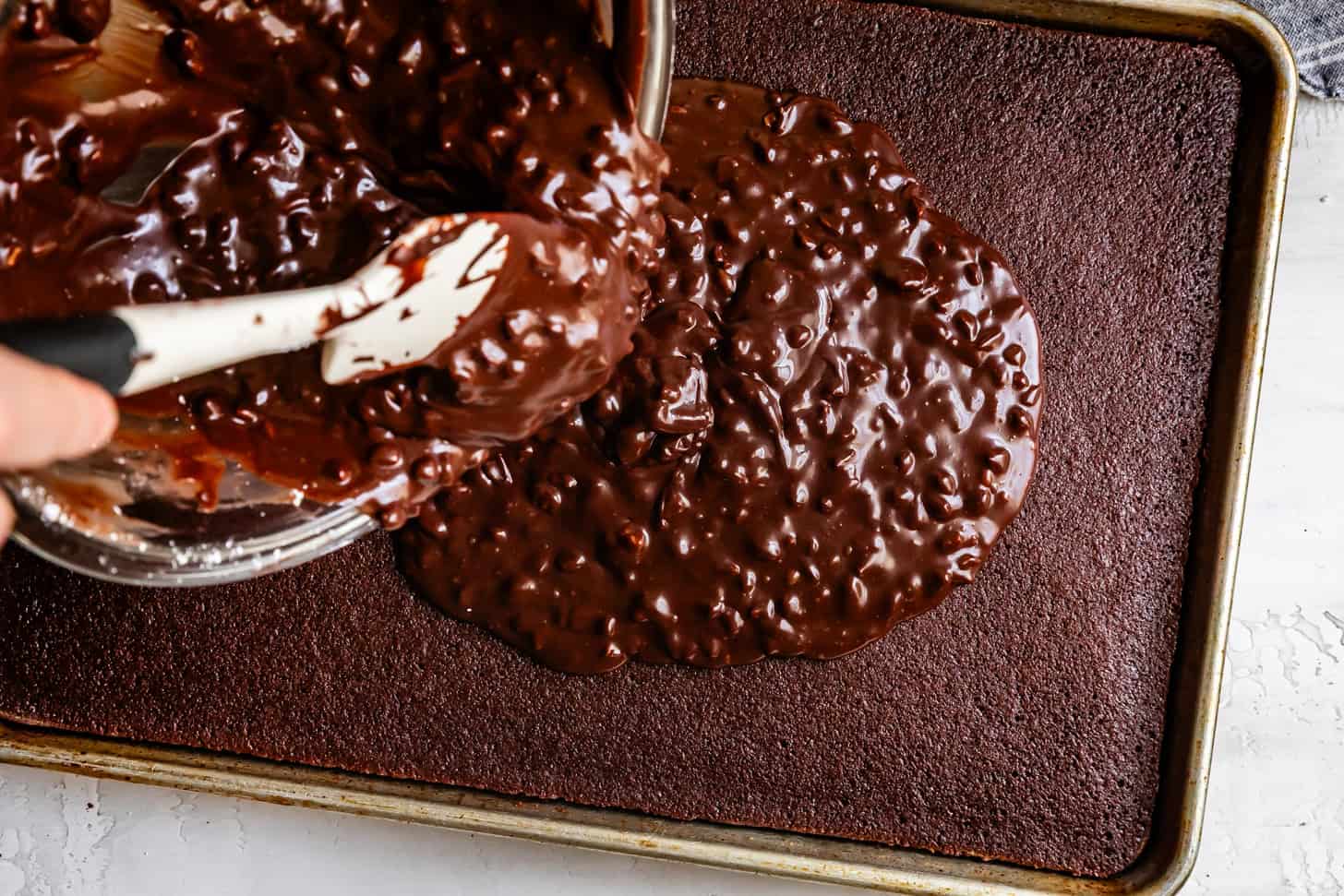 Pouring chocolate icing with pecans onto a Texas sheet cake in a baking pan.