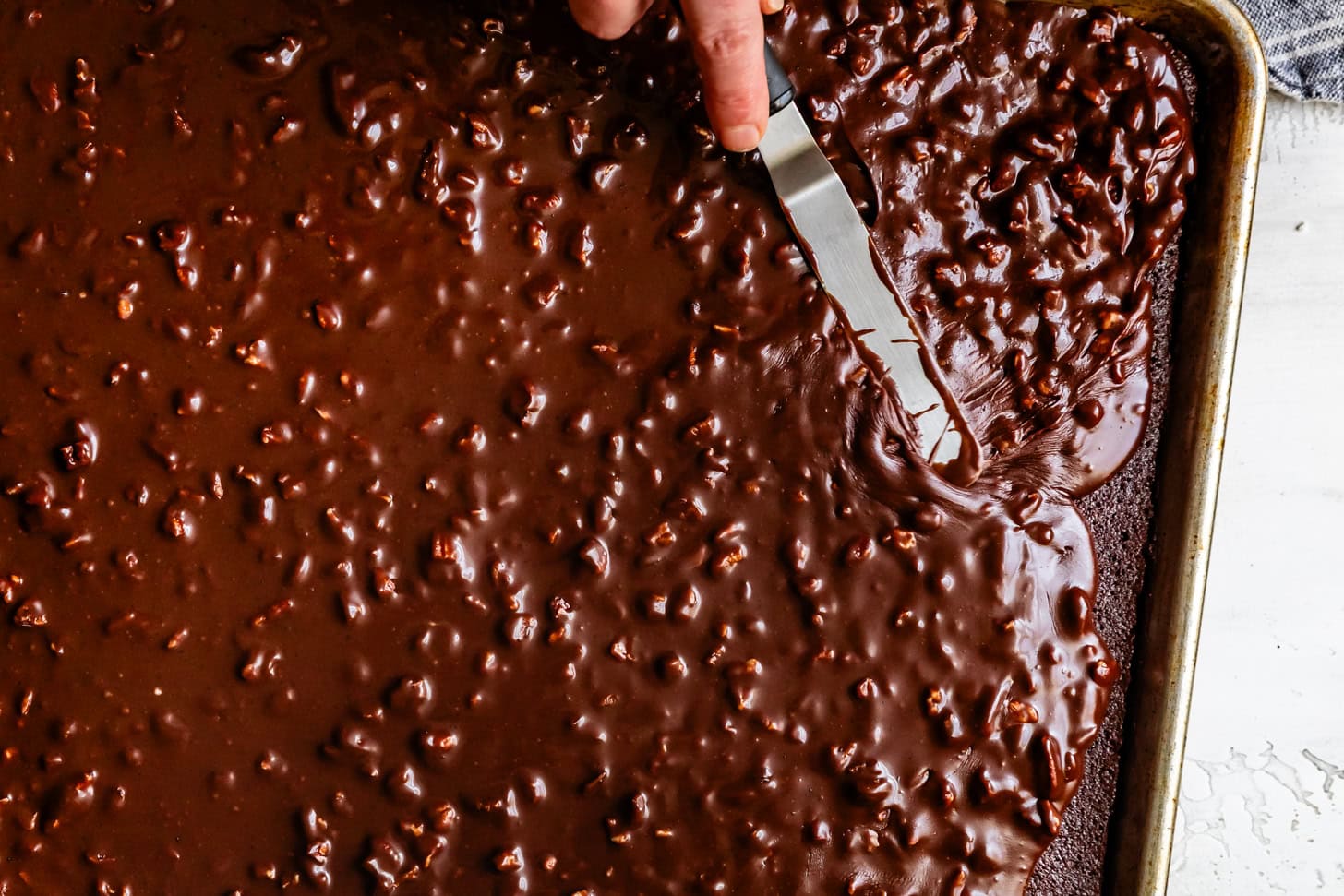 A hand spreads glossy chocolate icing with nuts over a gluten-free Texas Sheet Cake using an offset spatula.