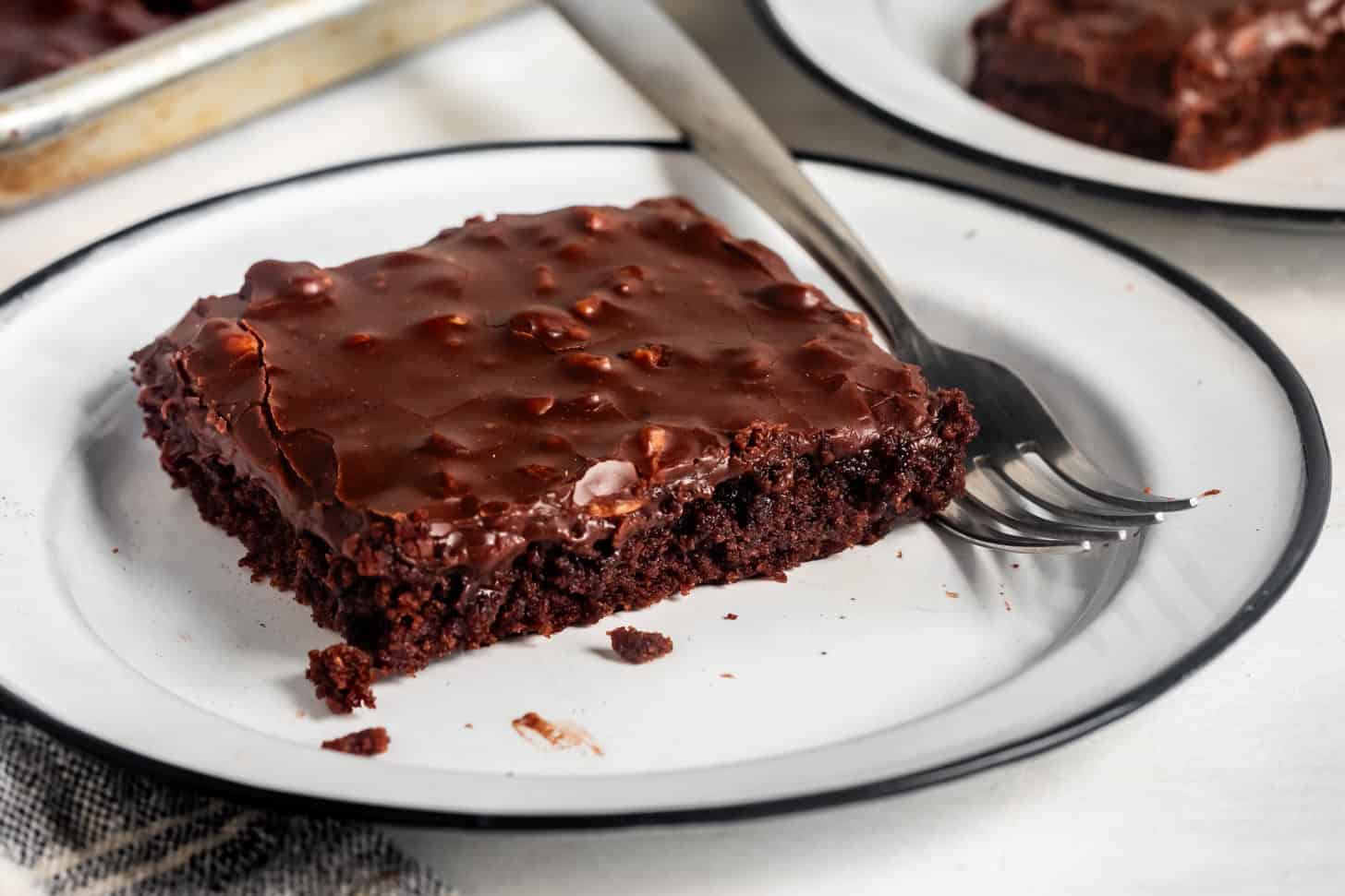 A square piece of fudgy chocolate gluten-free Texas sheet cake with rich chocolate icing sits on a white plate with a fork