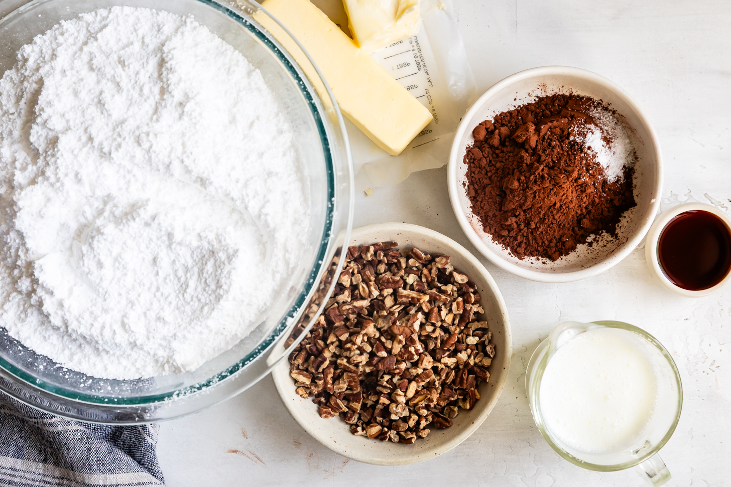 Bowls of powdered sugar, cocoa powder, chopped pecans, butter, milk, and vanilla on a white surface—perfect ingredients for creating a rich Fudgy Icing.