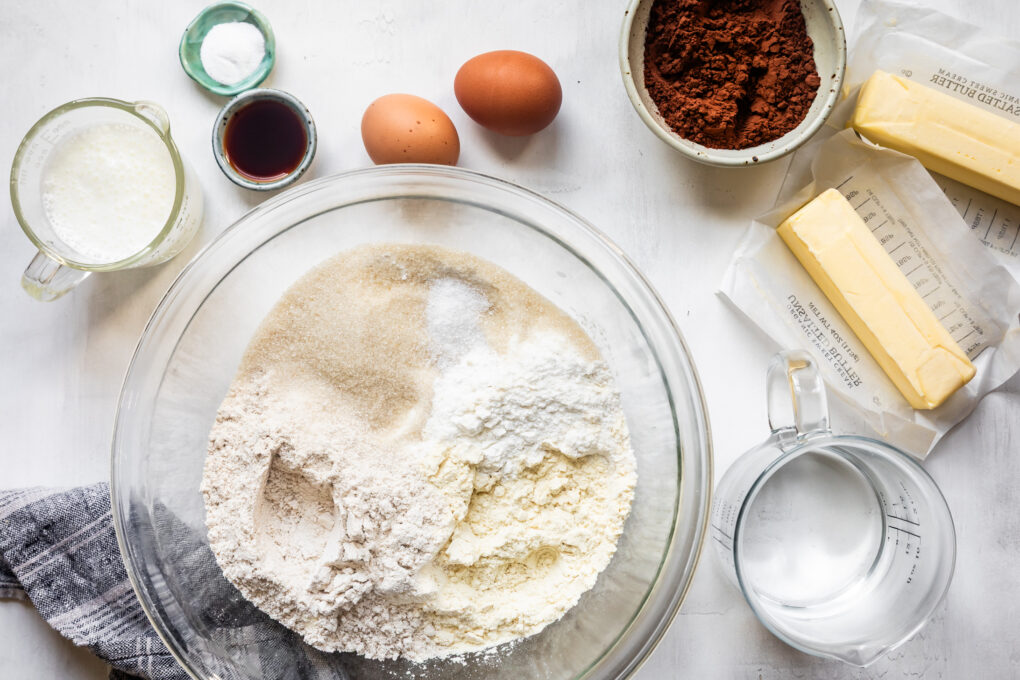 Baking ingredients on a white table: flour, sugar, eggs, butter, cocoa, milk, vanilla, water, and salt-everything you need for a classic Buttermilk Gluten-Free Texas Sheet Cake.