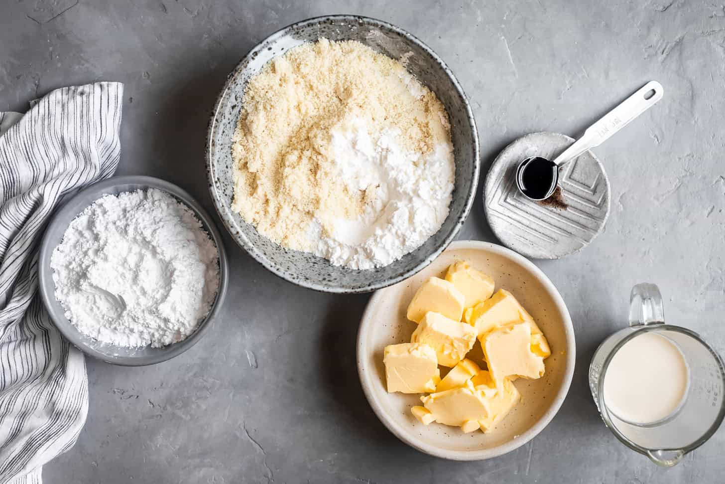 Bowls of almond flour, powdered sugar, butter, and a measuring cup of milk on a gray surface with a striped towel-perfect for making gluten-free butter cookies.