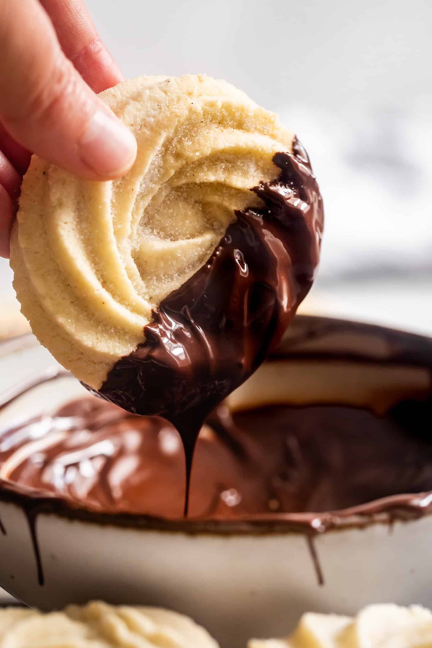 A hand dips a swirled, gluten-free butter cookie halfway into melted chocolate above a bowl.