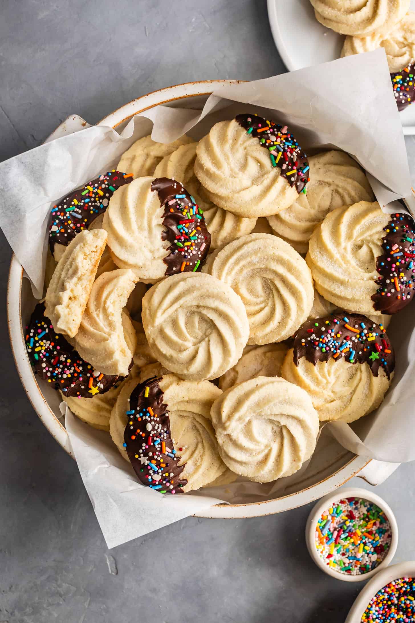 A bowl of swirled gluten-free butter cookies made with almond flour, some dipped in chocolate and sprinkles, with parchment paper.