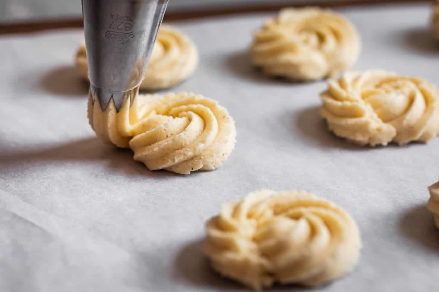 A piping bag forms swirled gluten-free butter cookie dough shapes on a baking sheet lined with parchment paper