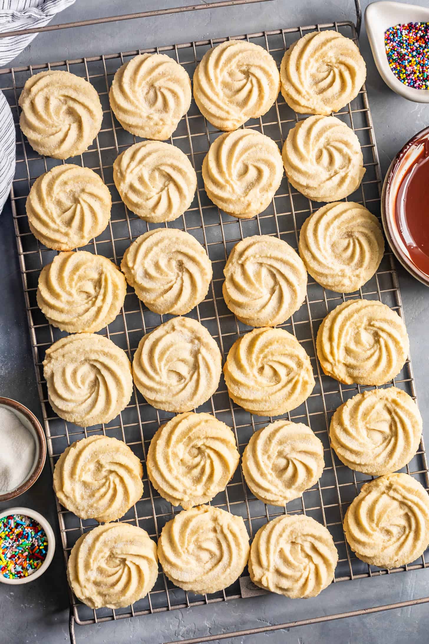 Swirled gluten-free butter cookies on a cooling rack, made with almond flour, with bowls of chocolate, sprinkles, and sugar nearby.
