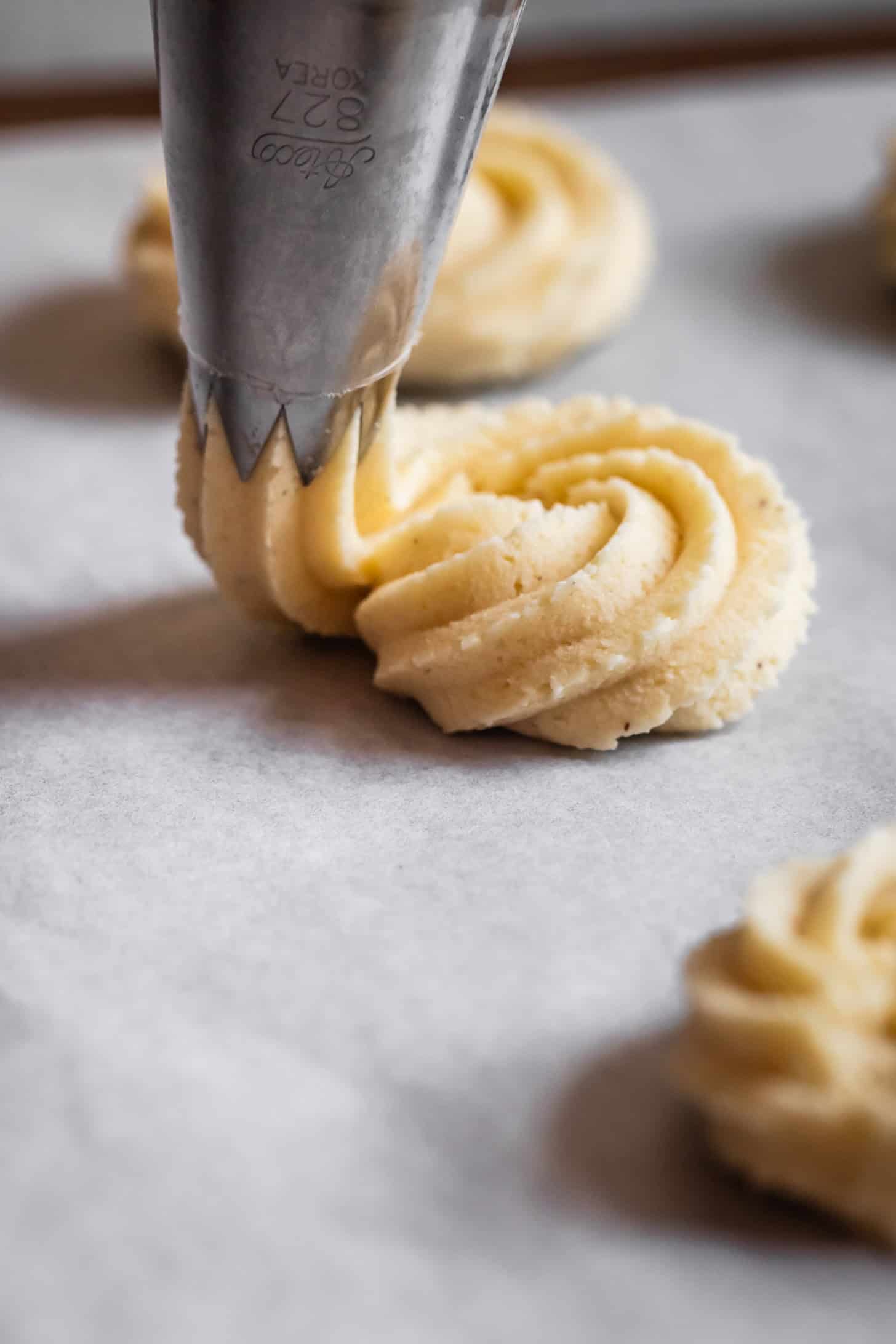 A piping bag squeezing gluten-free butter cookie dough onto parchment paper, forming a swirled shape before baking.